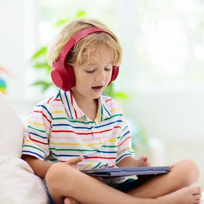 Child wearing red headphones and a striped shirt, holding a tablet representing young kids doing Lexia Core5 in short independant sessions anywhere.